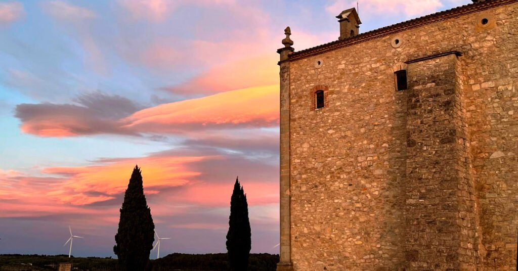 Fachada de piedra de una ermita histórica en La Fatarella al atardecer, con cipreses y aerogeneradores de fondo, en el corazón de Terra Alta