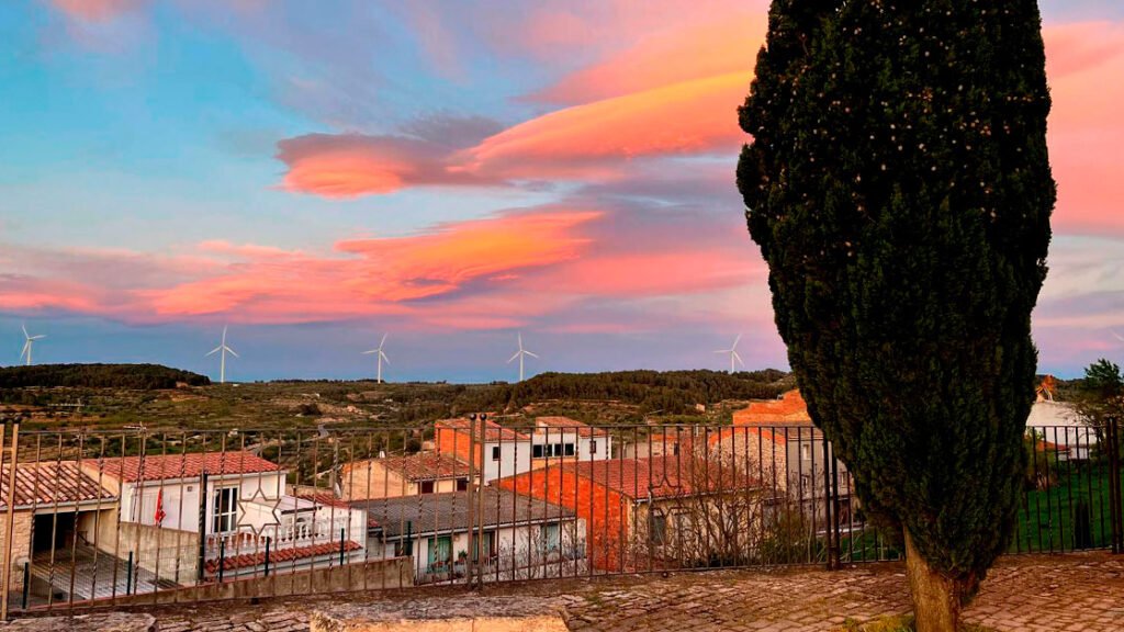 Vista panorámica de los tejados de La Fatarella al atardecer, con un ciprés, aerogeneradores y cielo rosado, en Terra Alta, Tarragona